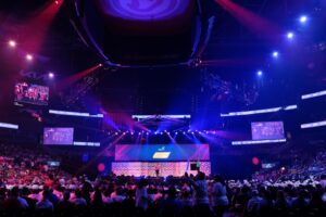 A vibrant stage setup at State Farm Arena for a large event, featuring dynamic lighting in purple and blue hues, multiple screens displaying live footage and graphics, and a packed audience seated below.