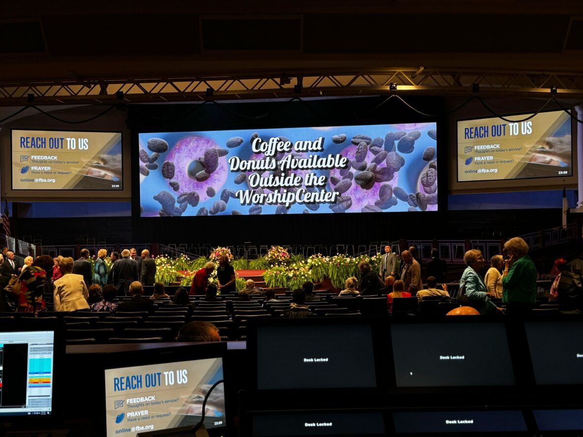 A large indoor event features a vibrant screen displaying the message "Coffee and Donuts Available Outside the Worship Center" along with two smaller screens on either side requesting feedback and prayer