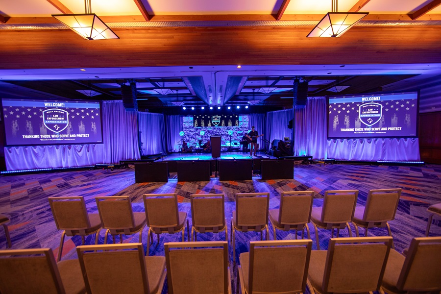 Ballroom event setup with a stage, multiple screens displaying a "Law Enforcement Appreciation" message, blue and purple lighting, and rows of chairs.