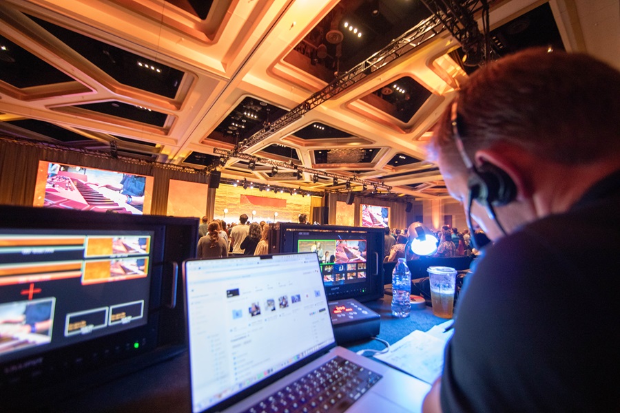 Event technician overseeing AV and lighting equipment setup during a corporate event, with multiple video screens and a control laptop visible in the foreground.