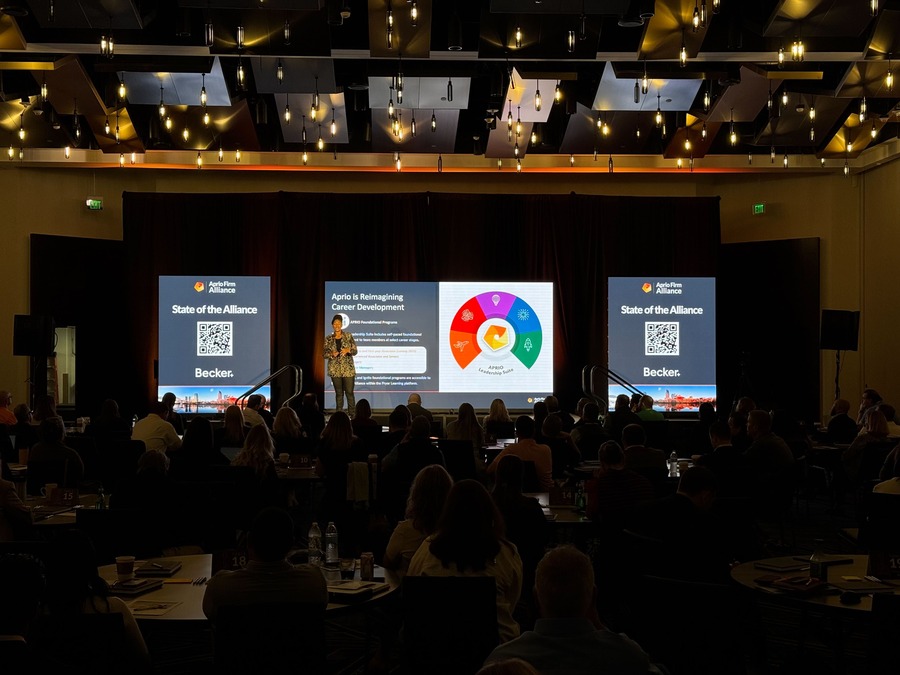 Conference stage with professional lighting, large LED screens showcasing 'State of the Alliance' visuals, and a presenter addressing an engaged audience in a modern venue.